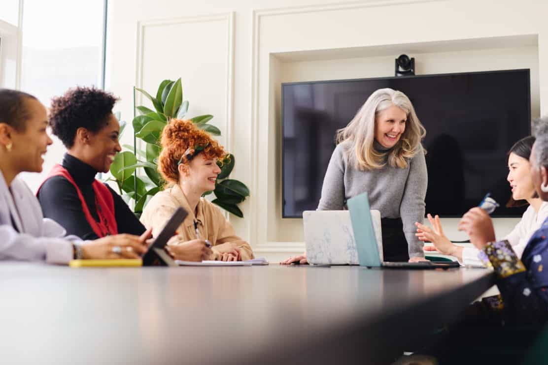 portrait-of-senior-white-businesswoman-with-laptop-smiling-and-listening-to-multiethnic-female-colleagues-in-boardroom