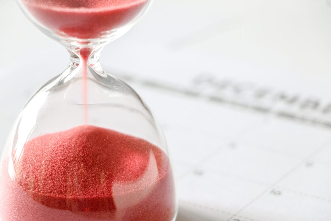 close-up-of-red-sand-slipping-away-through-hourglass-time-running-out-december-calendar-out-of-focus-in-background-end-of-year-no-more-time