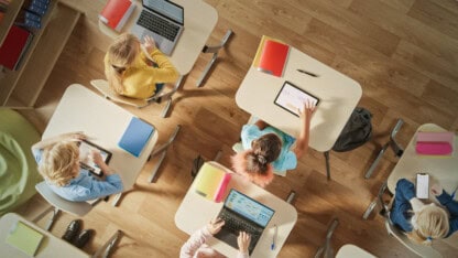 Overhead view of students at desks using laptops, tablets, and phones in a classroom.