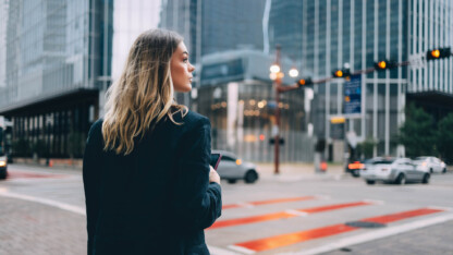 Person in business attire holding a smartphone at a downtown intersection, surrounded by modern office buildings and passing traffic.